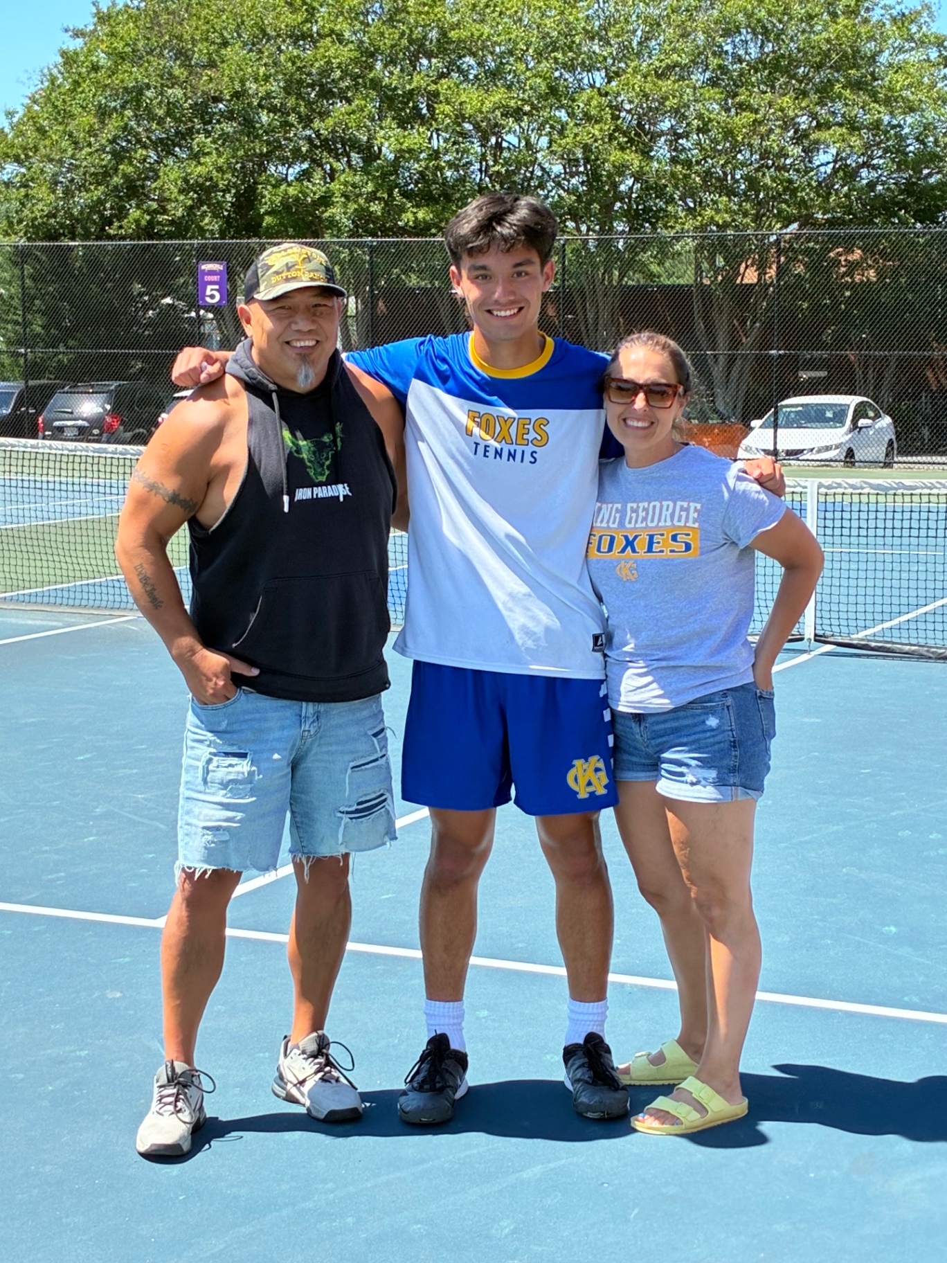 Judah Wang with his parents on the tennis court, wearing his KGHS Foxes tennis jersey
